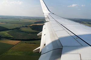 Airplane wing mid-flight above green fields under a blue sky, a Photo by AS photostudio