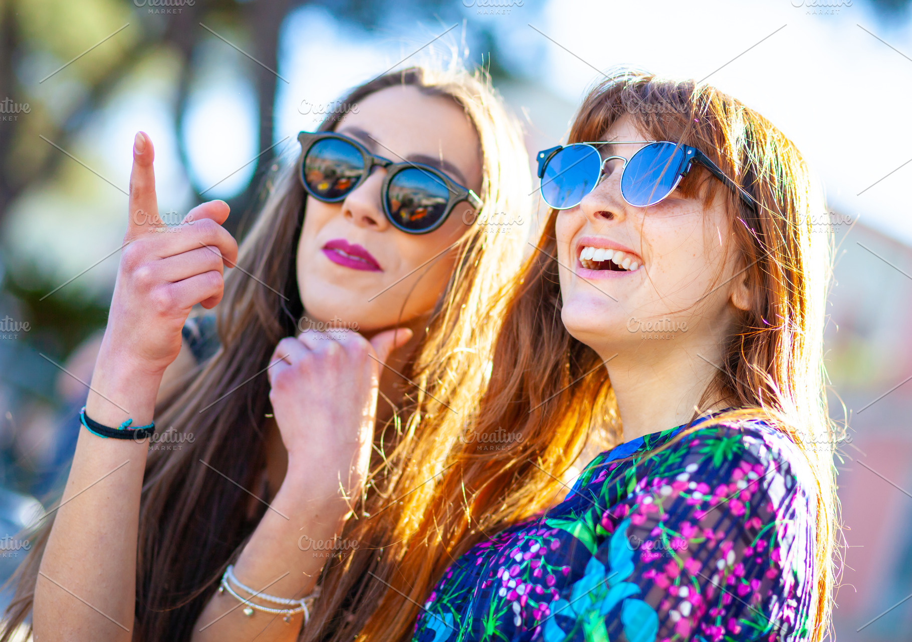 Two young women wearing sunglasses pointing and laughing outdoors, a Person Photo by Antonio Gravante Market