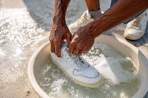 Hands cleaning a white sneaker in a bowl of soapy water by the beach on a sunny, a Photo by AlyaAnd