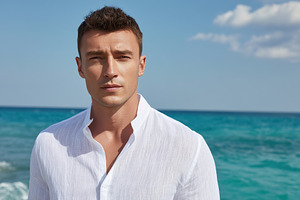 Man in white linen shirt stands by the ocean with a clear blue sky and waves in, a Person Photo by AlyaAnd