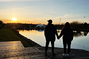 Couple enjoying a serene lakeside sunset while holding hands on a wooden deck, a Photo by AS photostudio
