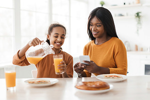 African Daughter Pouring Orange Juice In Glass For Mother Indoor, a Photo by Prostock-Studio