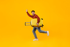 Happy Tourist Man Jumping Holding Suitcase Gesturing Yes, Yellow Background, a Photo by Prostock-Studio