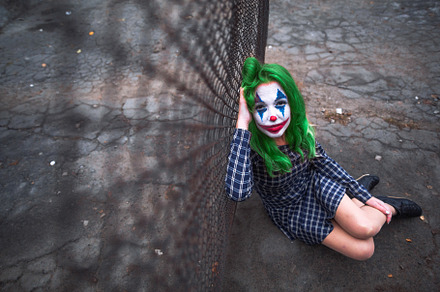 Greenhaired girl in chekered dress with joker makeup sitting near wire mesh fen