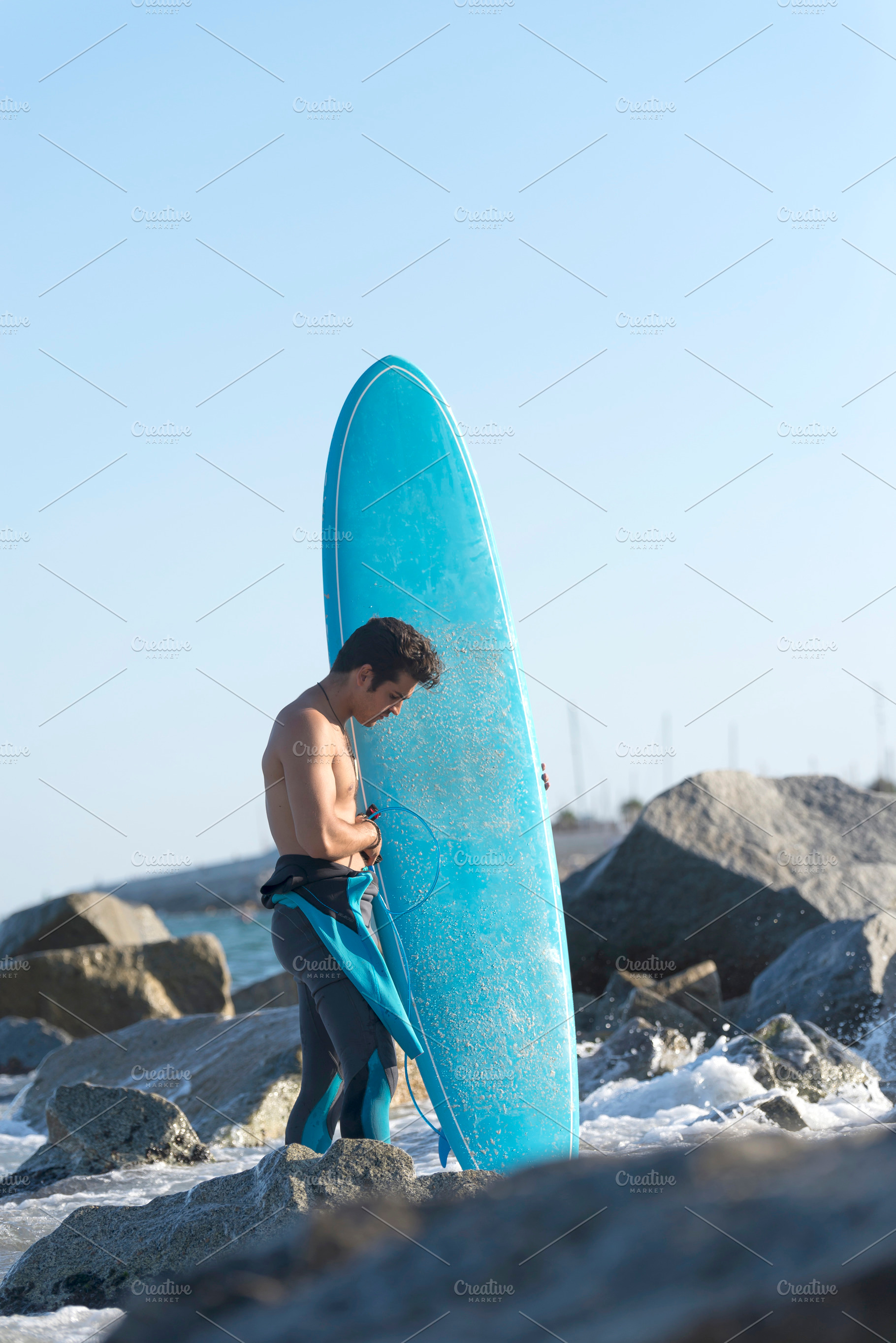 Back view of young surfer standing barefooted on sandy shore,, a Holiday Photo by 2Design