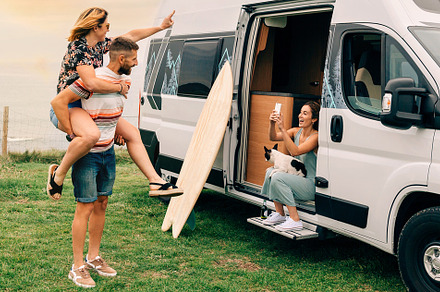 Woman taking photo with mobile of her friends piggybacking next to their camper, a Holiday Photo by davidpereiras