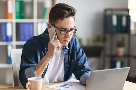 Young Man Working On Laptop And Talking On Mobile Phone In Office