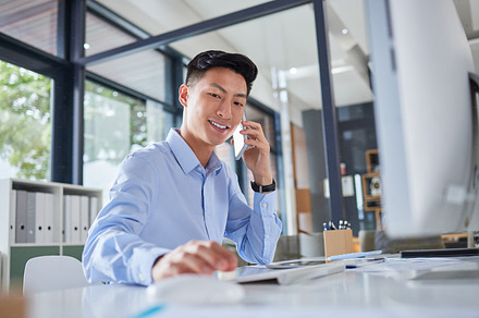 Young asian businessman making a call on his cellphone. Happy businessman typing on his computer. Business professional working in his office. Businessman calling on his smartphone., a Photo by Peopleimages.com