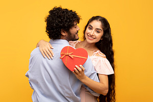 Young indian lovers hugging on yellow background, lady holding heart shaped gif, a Photo by Prostock-Studio