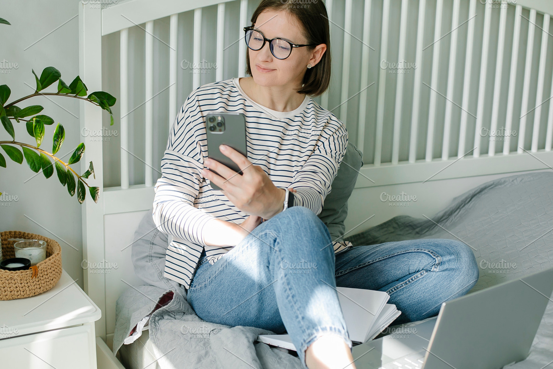 Beautiful young woman talking on phone, using laptop relaxing sitting on bed at, a Person Photo by Anikona
