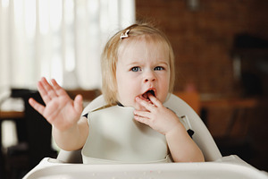 Infant girl or toddler sitting on high chair. Eating food by herself using spoo