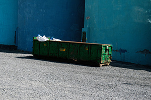 Green dumpster against blue walls in a sunlit area in Akranes, Iceland, a Photo by AS photostudio