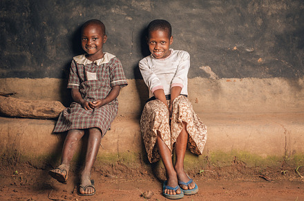 Two African Girls Sit & Smile at Hut