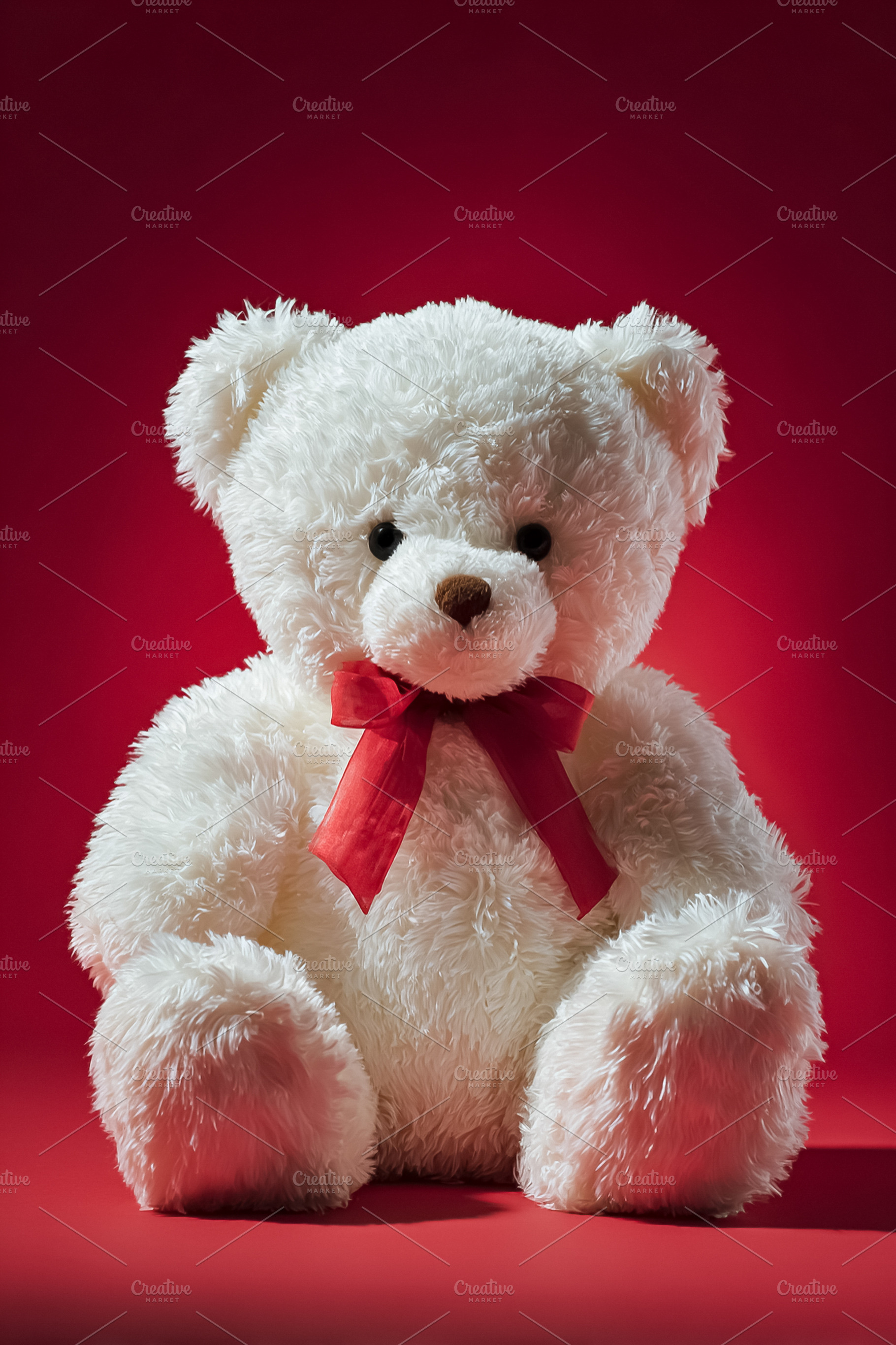 White teddy bear with red ribbon sitting on a vibrant red backgr, a Photo by Studio ANNA