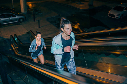 Women friends training running up escalator in city at night