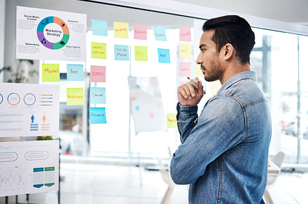 Creative man, thinking and glass board for project management, marketing or planning at office. Male person, manager or employee checking project plan, tasks or statistics for startup at workplace, a Photo by Peopleimages.com