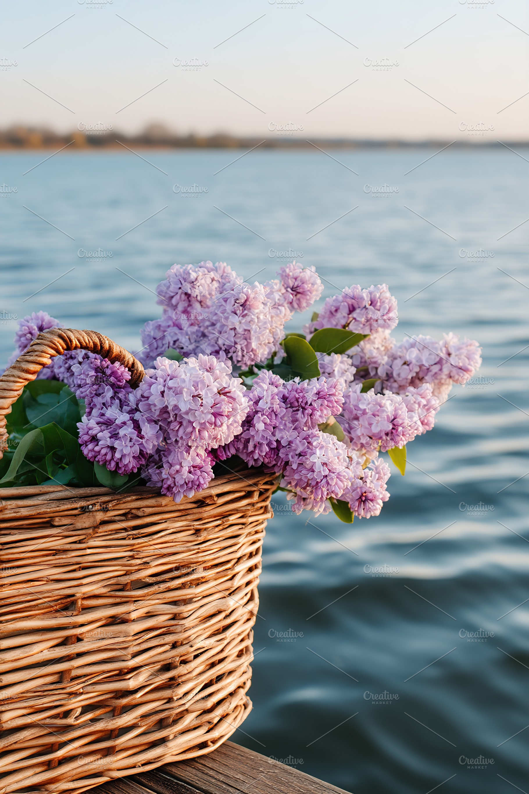 Fresh purple lilac flowers in a wicker basket by a calm lake during sunset, a Nature Photo by AlyaAnd