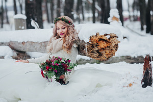 Cute bride sits on the snow near to the wood log and holds a bouquet winter