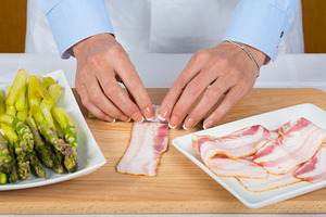 Chef wrapping asparagus with bacon slices containing asparagus, peeling, and