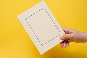 A person holding a closed light hardcover book with a frame on t, a Person Photo by ndanko