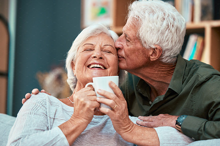 Coffee kiss, love and senior couple smile for retirement, conversation and relax in the living room of a house. Home tea, affection and elderly man and woman with comic communication in the lounge, a Photo by Peopleimages.com