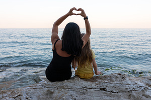 Back view of woman and child sitting on driftwood, making a heart shape with ha, a Person Photo by Ales Munt