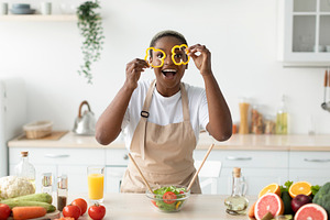 Glad young black female in apron have fun, make salad, puts pieces of paprika t, a Person Photo by Prostock-Studio
