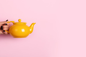 Female hand holding a bright yellow teapot against a pastel pink, a Holiday Photo by Ales Munt