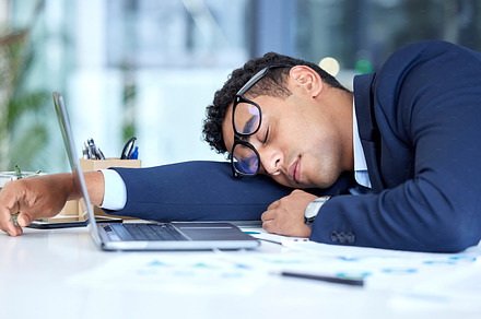 One exhausted mixed race businessman wearing glasses while sleeping at his desk in an office. Young entrepreneur feeling overworked, tired and demotivated. Lazy man slacking and ignoring deadlines by taking a nap. Burnout and stress in the workplace, a Photo by Peopleimages.com
