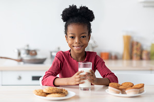 Cheerful Black Girl Sitting At Table, Enjoying Milk And Cookies, a Photo by Prostock-Studio