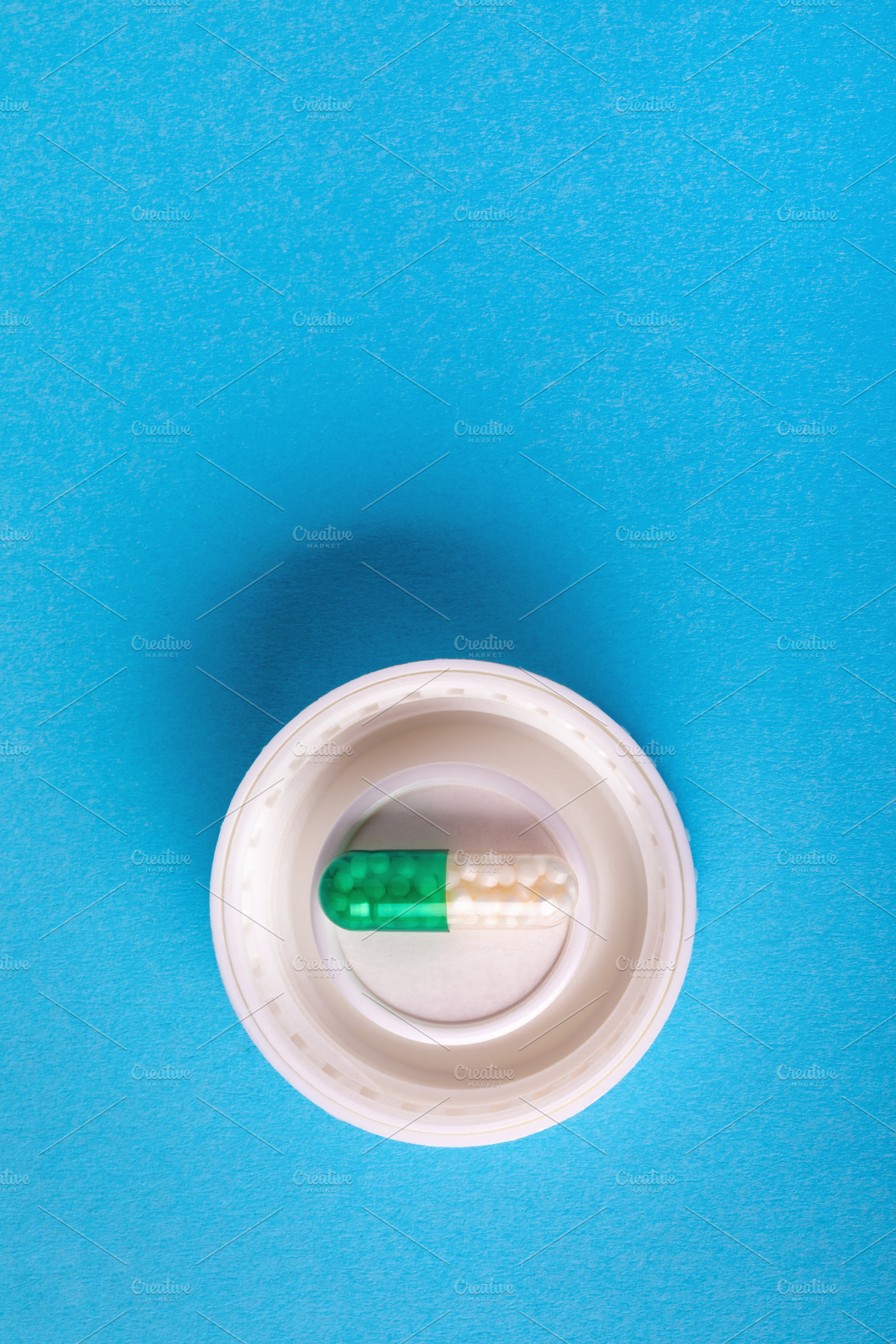 Green and white capsule pill placed inside a white bottle cap on a vibrant blue, a Health & Medical Photo by Ales Munt