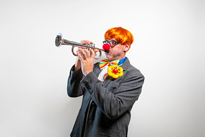 Clown playing trumpet with bright orange wig and red nose, a Person Photo by karrastock