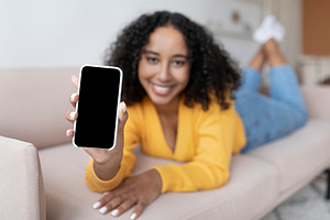 African American woman showing smartphone with blank screen, lying on sofa at h, a Photo by Prostock-Studio
