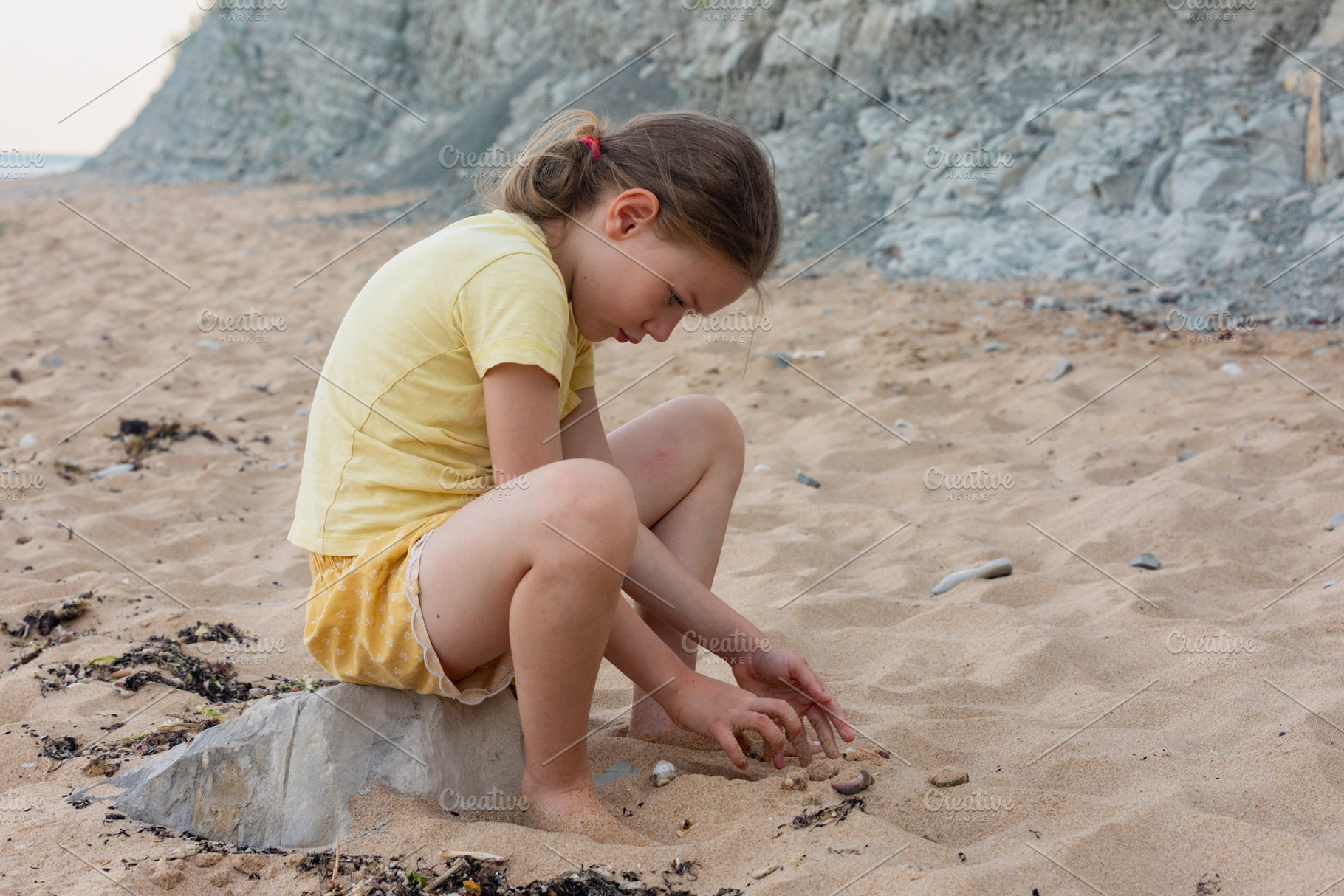 Young girl in yellow clothes playing with sand near rocky cliffs on a natural b, a Person Photo by Ales Munt