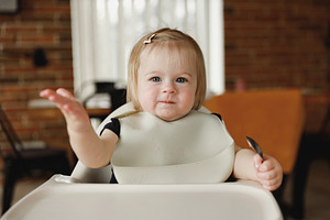 Infant girl or toddler sitting on high chair. Eating food by herself using spoo, a Person Photo by Lenor4ik