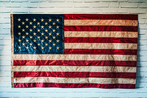Worn American flag hanging on white brick wall, a Background Photo by karrastock
