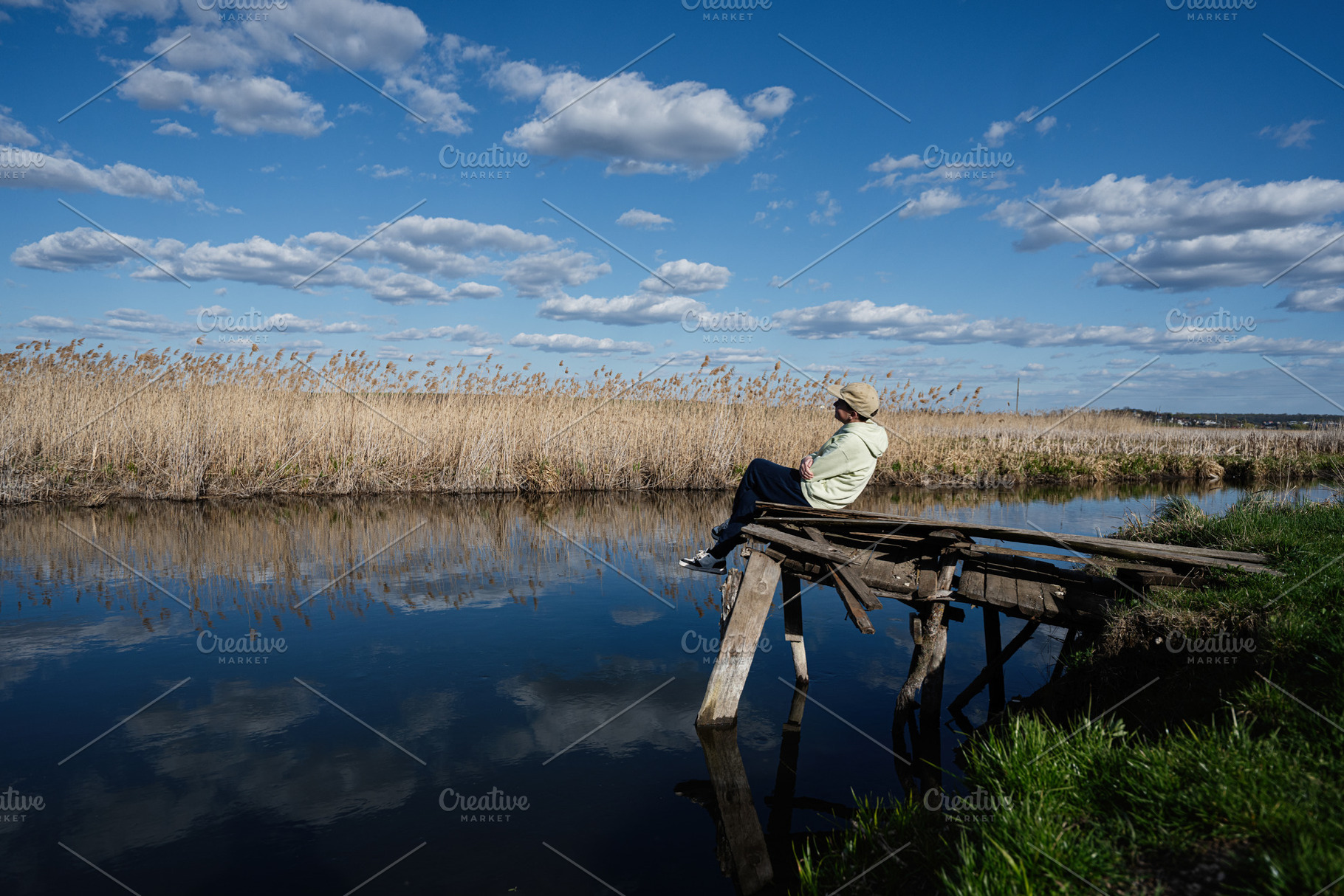Boy sitting on rustic wooden dock by river surrounded by reeds under blue sky w, a Person Photo by AS photostudio