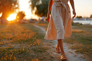 Woman in light linen dress and sandals walking along a scenic path during sunse, a Photo by AlyaAnd