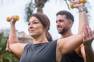Male personal trainer assisting woman with dumbbell exercise outdoors