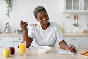 Happy millennial african american female in casual eating porridge, making note, a Person Photo by Prostock-Studio