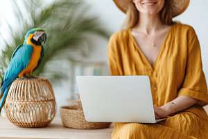 Colorful parrot sits on desk next to woman working on laptop in a bright enviro, a Person Photo by AlyaAnd