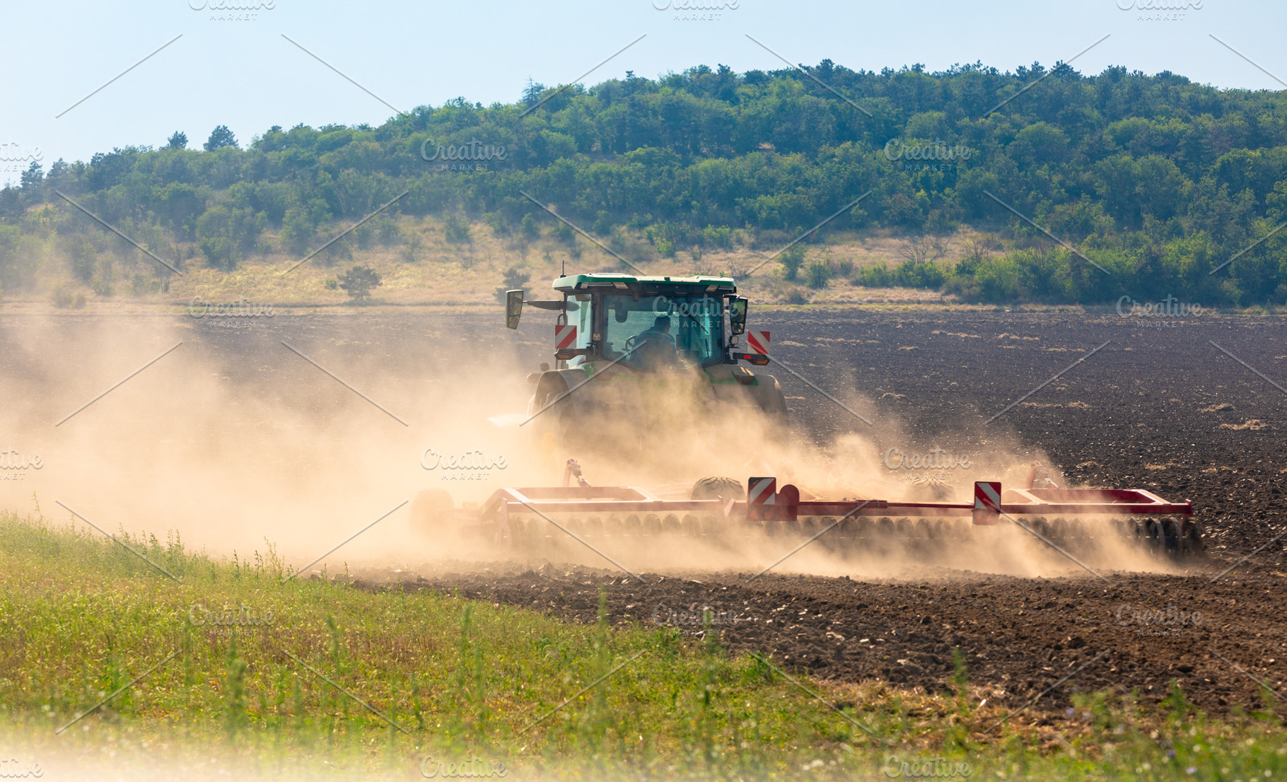 Modern tractor works on a sunlit field, creating dust clouds against a green fo, an Industrial Photo by Ales Munt