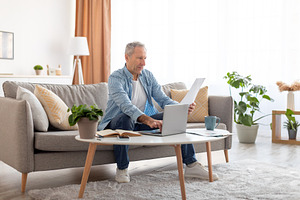 Man holding documents reading report working on laptop at home, a Photo by Prostock-Studio