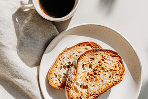 Enjoying coffee with toasted bread on a white ceramic plate in morning light, a Photo by AlyaAnd