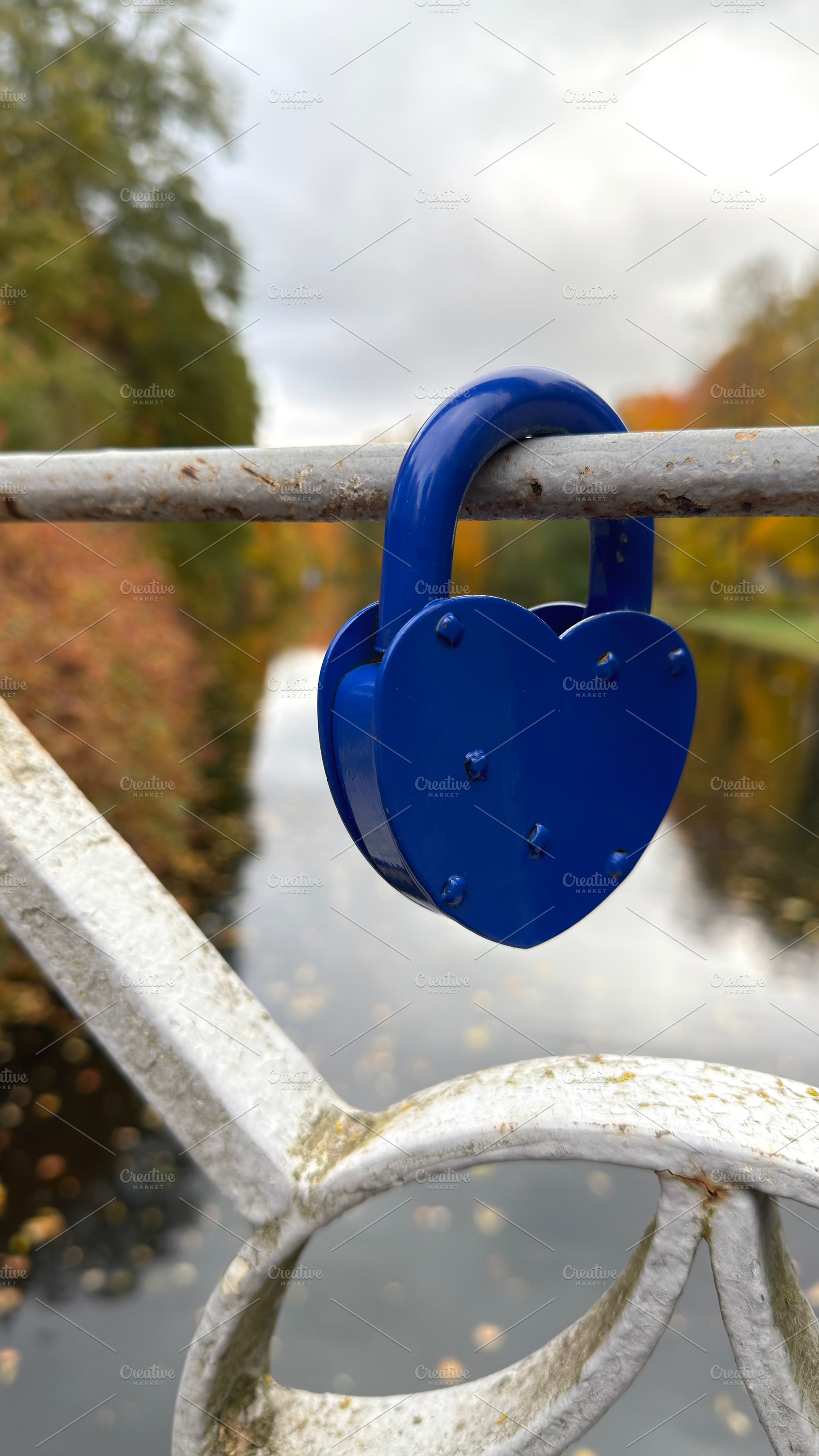 A blue heart-shaped padlock attached to a metal railing on a bridge, with a riv, an Industrial Photo by Textures & Backgrounds