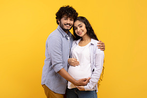 Satisfied young indian man hugging pregnant wife and touching belly, smiling to, a Photo by Prostock-Studio