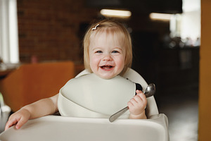 Infant girl or toddler sitting on high chair. Eating food by herself using spoo, a Person Photo by Lenor4ik
