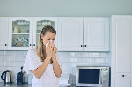 Sick caucasian woman holding tissue and blowing her nose while standing in kitchen at home. Woman suffering from seasonal allergy or flu, a Photo by Peopleimages.com