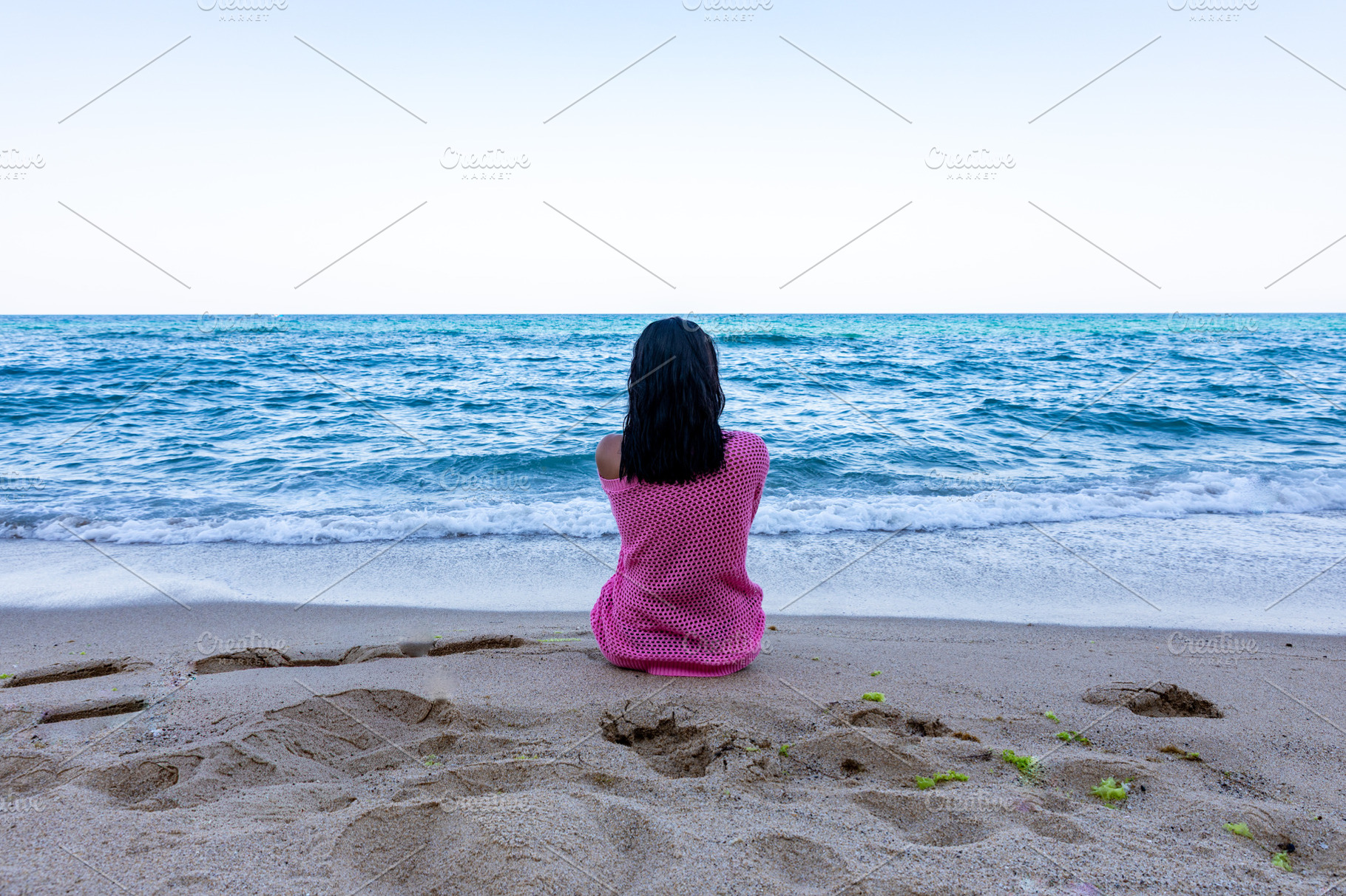 Rear view of a woman sitting alone on sandy beach facing the calm ocean under c, a Person Photo by Ales Munt